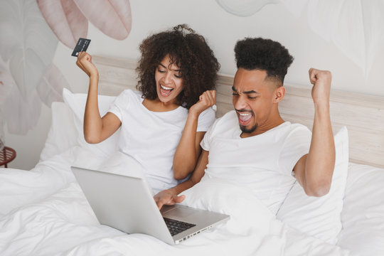 Young happy african loving couple girl guy in white t-shirts using laptop pc credit bank card lying on bed in bedroom at home, spending time in room. Rest relax good mood quarantine lifestyle concept.