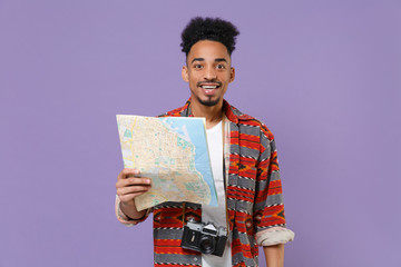Cheerful young african american guy in casual colorful shirt with photo camera on neck posing isolated on violet wall background studio. People lifestyle concept. Mock up copy space. Hold city map.