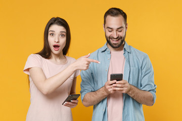 Shocked funny young couple friends guy girl in casual clothes isolated on yellow background. People lifestyle concept. Mock up copy space. Pointing index finger on mobile phone, typing sms message.