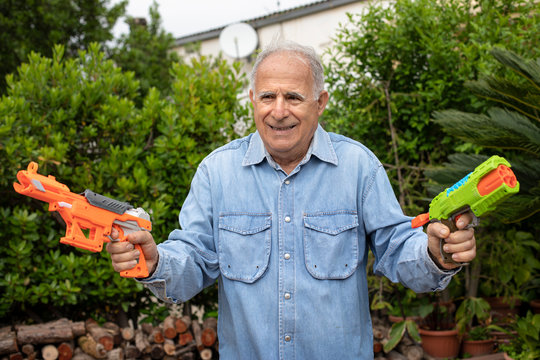 Anziano con camicia in jeans scherza allegramente nel suo giardino con due pistole ad acqua