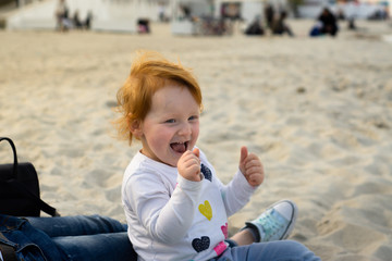 Cute red hair baby girl is seating on the beach near by her mothers knees smiling and making happy gestures with her hands. Sun set light scene