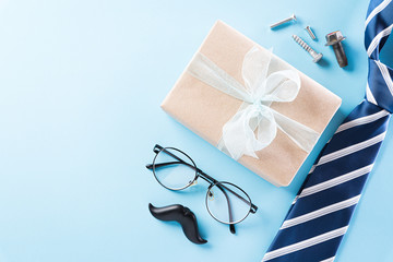 Happy Fathers Day background concept with blue necktie, glasses, black mustache and gift box on bright pastel background with copy space for text.