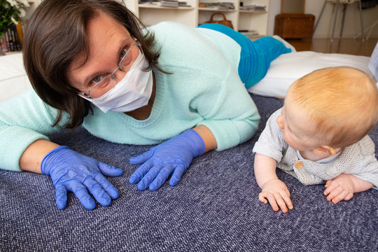 Babysitter Wearing Mask And Gloves Entertaining Baby At Home Interior, Playing With Child On Floor, Training To Crawl. Quarantine Or Babysitting Concept