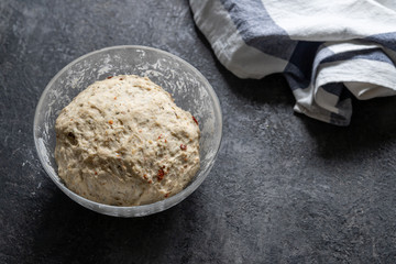 Knead yeast dough for homemade bread with herbs in a glass bowl next to a white-blue towel on dark table. Cook at home