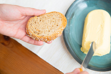 Woman cutting butter for slice of wholegrain freshly baked baguette. Closeup shot, high angle....