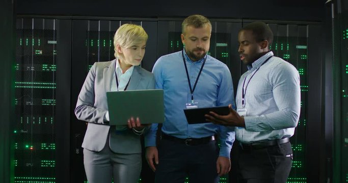 Multiethnic men and woman working at severs in analytic monitoring center and checking data on server with laptop and tablet. Consultation work in databace room with servers. Cybersecurity concept.