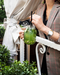 Cold kiwi lemonade and lime. Mug in the form of a jar. Drink with ice, green color. Against a background of flowers and greenery. The girl is holding a cocktail in her hand. Menu for cafes.Vegetarian.