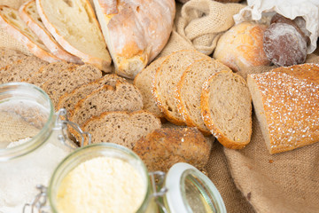 Slices, loafs and buns of traditional wheat and rye whole grain bread, burlap sackcloth and flour in jar. Top view. Bakery or traditional bread concept
