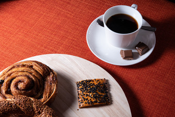 Cup of coffee with chocolate on the side. Crispy brown baked biscuits with black sesame seeds on wooden board. Coffee break bites. Bright red textile backdrop for copy space.