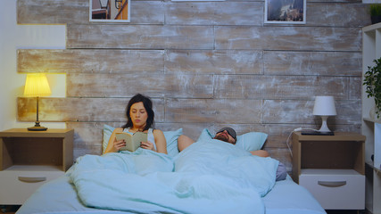 Beautiful young wife reading a book wearing pajamas while the husband is covering the face with...