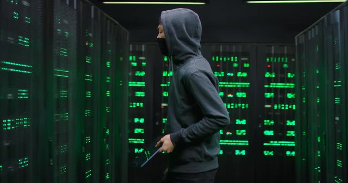 Asian male hacker in hood spying in server room with tablet device in hands and breaking into database system. Man, cyber crominal looking for passwords to hardware.