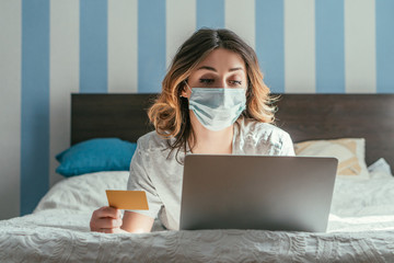 woman in medical mask holding credit card near laptop at home