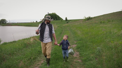 Dad with a fishing rod on his shoulder holding the hand of a little son with a bucket go fishing along the lake