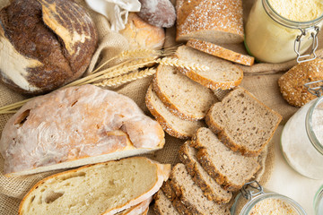 Table with wheat and rye whole grain loafs and baguettes cut in slices, corn flour in jar and ears. Top view. Bakery or traditional bread concept