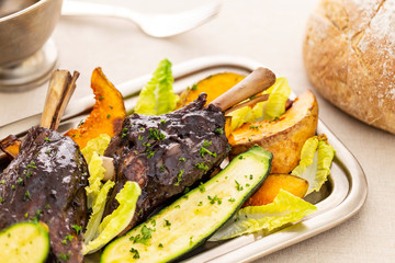 Glazed braised lamb shank with large pieces of pumpkin and zucchini on a silver tray on a beige tablecloth aside a vintage silver sauce boat and a bread loaf.