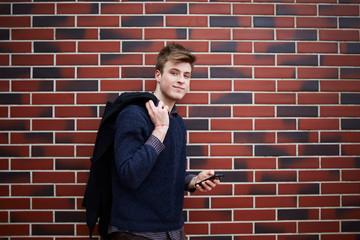 Fototapeta na wymiar Young man standing at brick wall with smartphone in hand