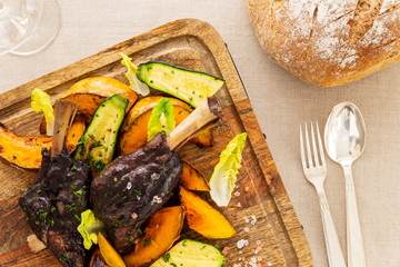 Glazed braised lamb shank with pumpkin and zucchini on a chopping board on a beige tablecloth aside a fork, a spoon, a bread loaf and a wine glass. Flat lay top view.