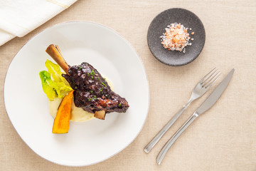 Glazed braised lamb shank on mashed potatoes with pumpkin and salad on a beige tablecloth aside fork, knife and a stone plate with pink Himalayan salt. Flat lay top view.