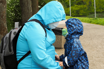 father and son in the Park during quarantine. a young man in a blue jacket with a gray backpack is sitting on a bench and a small four-year-old boy in a camouflage jacket is standing next to him