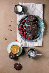 Homemade dark chocolate salted brownies cookies decorated by dry and fresh raspberries, served with salt flakes, berries, mint, milk and cup of coffee over brown texture background. Flat lay, space