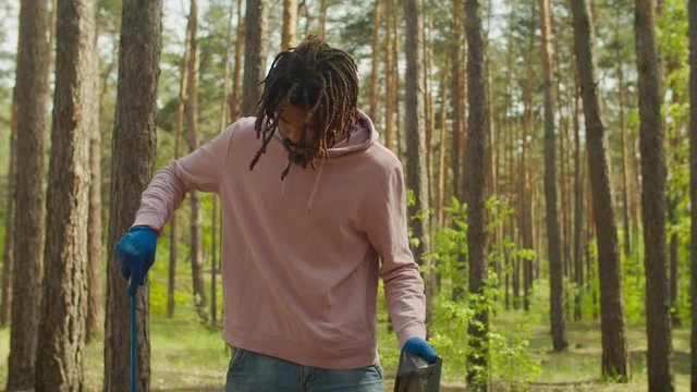 Handsome african man volunteer in casual clothes wearing protective gloves, picking up garbage and plastics using garden tool during cleaning forest, caring and preserving ecology.