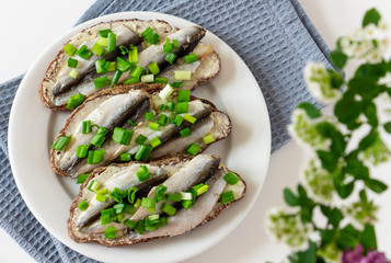 composition of food and flowers on a white background, three delicious sandwiches with rye bread, butter, small salted fish and green onions on a white plate, flower - white spirea