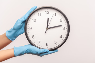 Profile side view closeup of human hand in blue surgical gloves holding analog clock. indoor,...