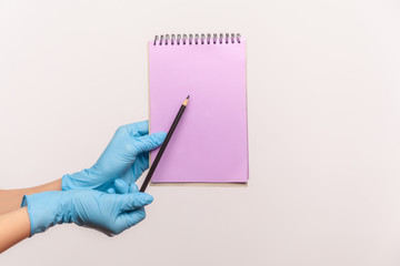Profile side view closeup of human hand in blue surgical gloves holding purple notepad in hand and holding empty paper with pencil. indoor, studio shot, isolated on gray background.