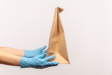 Profile side view closeup of human hand in blue surgical gloves holding and craft paper packet with food. safe food delivery concept. indoor, studio shot, isolated on gray background.