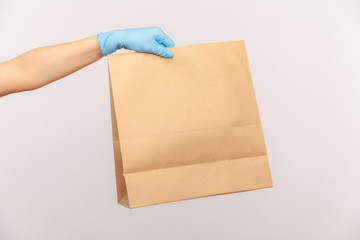 Profile side view closeup of human hand in blue surgical gloves holding and craft paper packet with food. safe food delivery concept. indoor, studio shot, isolated on gray background.