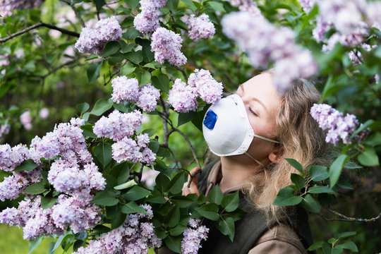Young blonde woman smells lilac tree blossom through FFP2 respirator. Conceptual image of corona virus quarantine and allergy