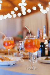 Banquet. Cover the table with a white tablecloth. On the table are glasses of aperol spritz with a straw for cocktails, dishes with food. Against bokeh