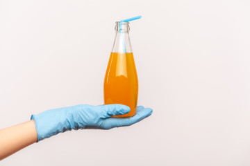 Profile side view closeup of human hand in blue surgical gloves holding bottle of orange drink. orange juice or cocktail in hand. safe food concept. indoor, studio shot, isolated on gray background.