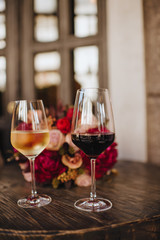 Two glasses of white and red wine stand on a table. Nearby is a bouquet of red roses