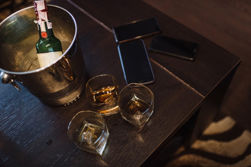 On a dark brown wooden table are three glasses of whiskey, a smartphone, an ice bucket and a bottle of whiskey.