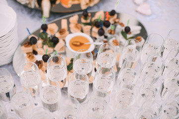 Many glasses with champagne are on the table. The table is covered with a white tablecloth. Nearby is a tray with canapes