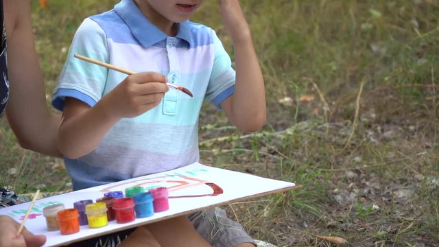 Closeup view video footage of cute little boy of 5 years old painting outdoors together with his mommy sitting together on ground outdoor in summer park. 