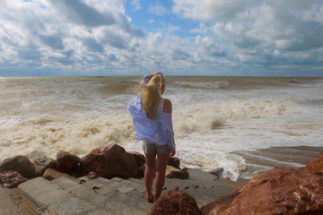 A young woman in the blue tunic and shorts is standing on the summer beach and looking at the stormy spindrift waves and clouds. View from the back. Stormy weather on the sea coast. Stormy summer day.