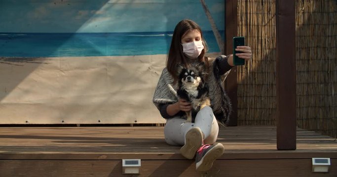 A teen age girl makes selfie while wearing protecting mask. She tries to make a photo with her dog. Self isolation in countryside while dreaming about sunny beach. Sunny backyard.
