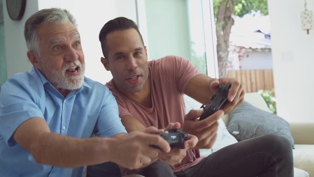 Senior Hispanic father with adult son sitting on sofa at home playing video game together - shot in slow motion