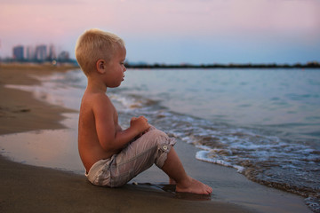 Fototapeta na wymiar Little baby boy sitting on the beach near sea and looking on the waves, boy on the seaside in sunset