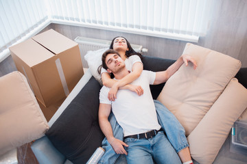 Couple resting on couch after moving in, man and woman relaxing on sofa just moved into apartment with cardboard boxes on floor, happy satisfied homeowners enjoying first day in new home, top view