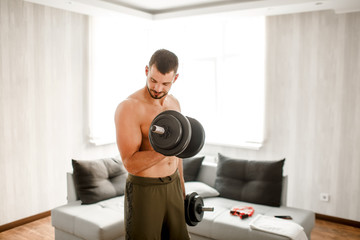 Young ordinary man go in for sport at home. View of hardwoking t-shirtless guy sportsman stand on floor and hold dumbbells in hand. Ready to start workout. Ordinary man work on improving his body.