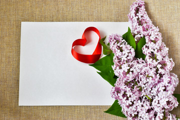 A bouquet of lilacs and a sheet of paper for writing congratulations and a heart made of ribbon, on a beige background. Holiday card. Holiday concept. Place for text.