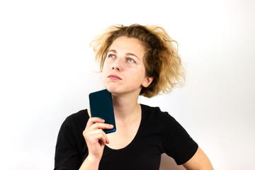 Fototapeta premium A girl with yellow hair holds a smartphone next to her face and looks up thoughtfully against a white background. Modern technologies, youth, emotions