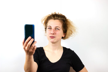 Naklejka premium A girl with yellow hair is talking on a video link on a smartphone and looks up in surprise, questioningly, dreamily, against a white background. Modern technologies, youth, emotions