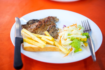 Dish of beef steak with salad, french fries and bread on red background