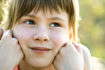 Portrait of pretty child girl with gray eyes and long fair hair leaning on her hands smiling happily outdoors on blurred bright background. Cute female kid on warm summer day outside.