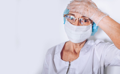 Elderly tired exhausted mature woman doctor or nurse holding head in a white medical coat, gloves, face mask wearing personal protective equipment. Healthcare and medicine. Covid-19 pandemic crisis