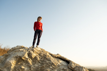 Young woman hiker standing alone on big stone in morning mountains. Female tourist on high rock in wild nature. Tourism, traveling and healthy lifestyle concept.
